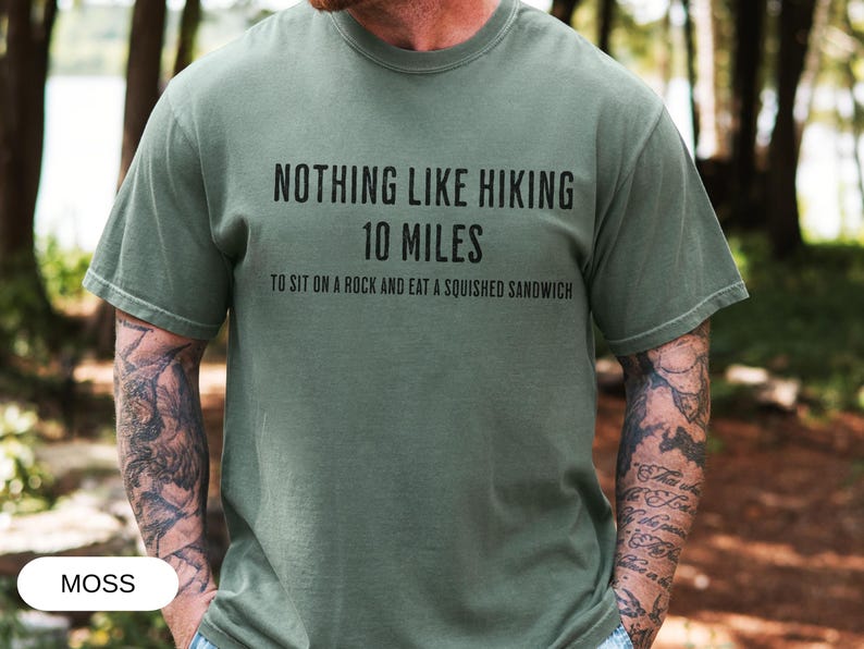 May include: Moss green t-shirt with the text "NOTHING LIKE HIKING 10 MILES TO SIT ON A ROCK AND EAT A SQUISHED SANDWICH" in black. The shirt is a casual style, suitable for outdoor activities.
