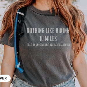 May include: Dark gray t-shirt with the text "NOTHING LIKE HIKING 10 MILES TO SIT ON A ROCK AND EAT A SQUISHED SANDWICH" in white. The shirt is worn with a blue hydration pack. The word "PEPPER" is at the bottom.