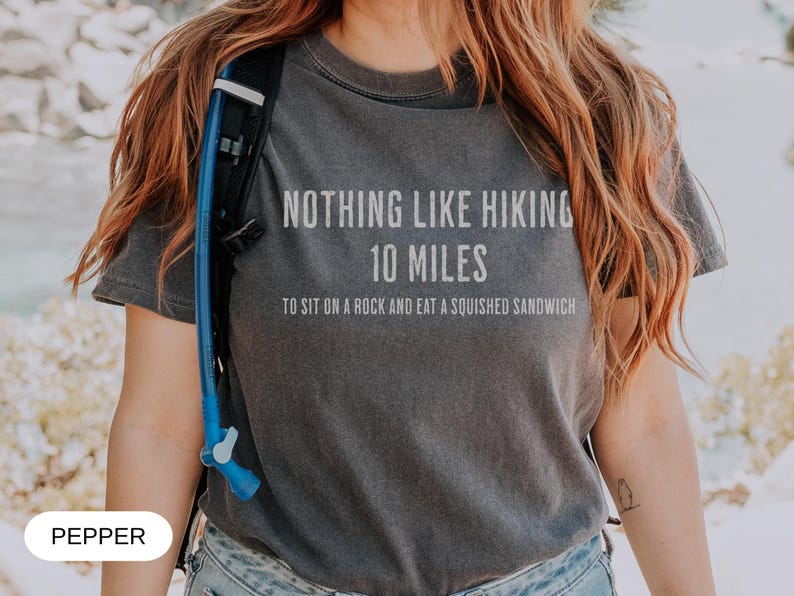May include: Dark gray t-shirt with the text "NOTHING LIKE HIKING 10 MILES TO SIT ON A ROCK AND EAT A SQUISHED SANDWICH" in white. The shirt is worn with a blue hydration pack. The word "PEPPER" is at the bottom.