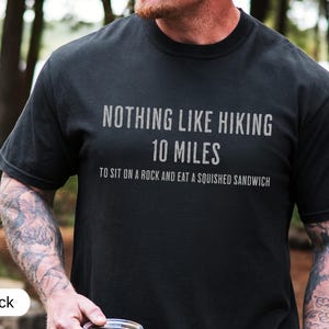 May include: Black t-shirt with the text "NOTHING LIKE HIKING 10 MILES TO SIT ON A ROCK AND EAT A SQUISHED SANDWICH" in a gray font. The shirt is made of a soft material and is suitable for outdoor activities.