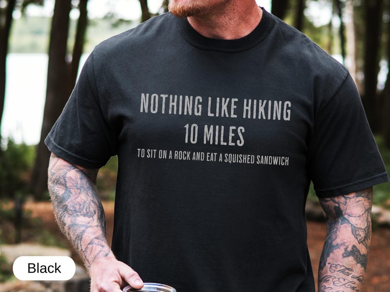 May include: Black t-shirt with the text "NOTHING LIKE HIKING 10 MILES TO SIT ON A ROCK AND EAT A SQUISHED SANDWICH" in a gray font. The shirt is made of a soft material and is suitable for outdoor activities.