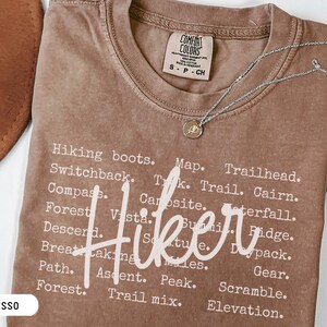 May include: Espresso-colored t-shirt with the word "Hiker" in a cursive font. The shirt features a list of hiking-related words in a white font. A silver necklace with a pendant is on the shirt.