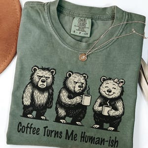 May include: Sage green t-shirt with a graphic of three bears, each holding a coffee mug. The bears have varying expressions, and the text "Coffee Turns Me Human-ish" is printed below. A gold necklace with a pendant is draped across the shirt.