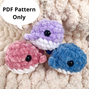 May include: Three crocheted dolphin keychains in pink, purple, and blue. The keychains are made with a fluffy yarn and have black eyes. The text "PDF Pattern Only" is in a speech bubble in the top left corner of the image.