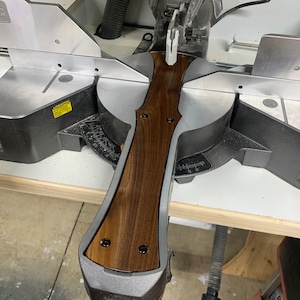 May include: A miter saw with a dark wood handle. The handle is attached to the saw's blade guard. The saw is silver and black, and the handle is a rich brown color. The saw is on a white surface.