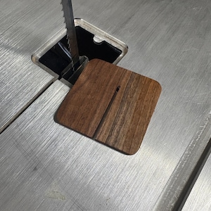 May include: A square, wooden band saw insert, made of dark brown wood, sits on a metal band saw table. The insert has a thin, vertical slot for the saw blade. The saw blade is visible in the image.