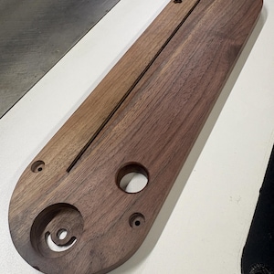 May include: A wooden table saw insert with a dark brown finish. The insert has a long, rectangular shape with rounded ends and a central groove. It features several pre-drilled holes and a circular cutout.