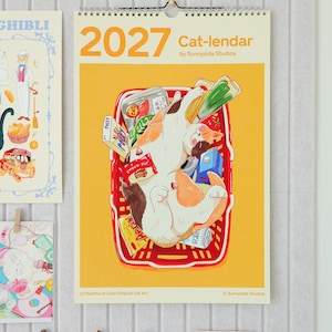 May include: A 2027 wall calendar titled Cat-lendar by Sunnyside Studios featuring a yellow background and a central illustration of a calico cat sleeping inside a red plastic shopping basket filled with various food items and snack packages.