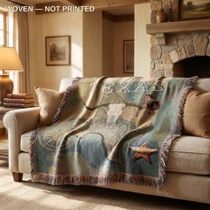 Peut inclure: Plaid tissé représentant une carte du Texas avec un crâne de longhorn, une étoile et le mot "TEXAS". La couverture a une bordure à franges et est drapée sur un canapé beige. La palette de couleurs comprend des nuances de bleu, de beige et de marron.