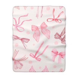 May include: A soft white blanket featuring a repeating pattern of various pink bows and ribbons. The design includes watercolor-style illustrations of bows in solid pink, striped pink, and checkered pink patterns, interspersed with small pink dots.