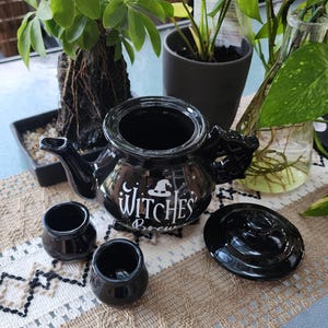 May include: Black ceramic teapot with a witch's hat and the words "Witches Brew" in white lettering. The teapot is accompanied by two small black ceramic cups with lids.