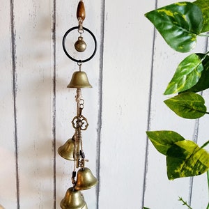 May include: A decorative wind chime with brass bells of different sizes. The chime includes a wooden bead, a black metal ring, and key charms. The bells hang from a natural twine cord, set against a white wooden backdrop.