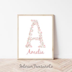 May include: A framed print featuring the letter "A" composed of pink floral elements. The name "Amelia" is written in a matching pink script below the letter. The print is in a light wooden frame, with the text "Interior Treasure Co" at the bottom.