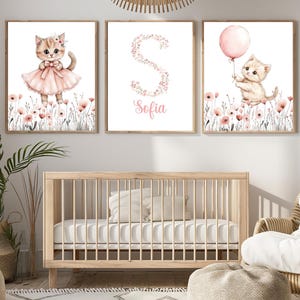 Puede incluir: Tres impresiones artísticas enmarcadas con ilustraciones de acuarela de gatitos. Un gatito lleva un vestido rosa, otro sostiene un globo rosa y el tercero muestra la letra "S" con el nombre "Sofia". Las impresiones están en un entorno de guardería.