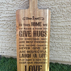 May include: A wooden cutting board with a handle, featuring a burnt-in message that reads "In this home we say please & thank you give hugs try everything once are best friends give second chances never give up are respectful laugh (a lot) play hard & have fun keep our promises love".
