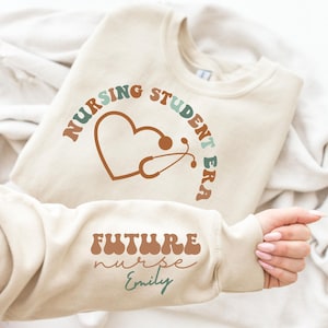 Puede incluir: Una sudadera beige con el texto "Nursing Student Era" en un arcoíris de colores, un corazón con un estetoscopio y el texto "Future Nurse Emily" en la manga.