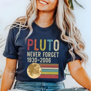 May include: A navy blue t-shirt with a vintage-style graphic that says "Pluto Never Forget 1930-2006". The graphic features a colorful rainbow stripe design with a yellow moon.
