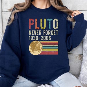 May include: A navy blue sweatshirt with a vintage-style graphic that says "PLUTO NEVER FORGET 1930-2006". The graphic features a yellow moon with a retro rainbow stripe design.