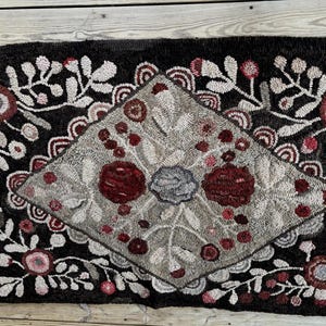 May include: A rectangular hooked rug featuring a dark brown background with a central diamond shape in light gray. The design includes floral and berry motifs in shades of deep red, light pink, and gray, with intricate white leaf patterns throughout.