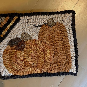 May include: A rectangular rug featuring a hooked fiber design of two orange pumpkins against a cream-colored background. The piece is framed by a thick black border with a small section of yellow and black striped pattern in the top left corner.