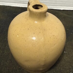 May include: A tan-colored, round jug with a small handle and a narrow opening. The jug has a speckled surface and a smooth, glossy finish. The jug is likely made of ceramic or stoneware.