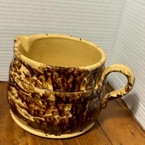May include: A vintage, handmade ceramic pitcher with a tan interior and exterior. The exterior features a dark brown, mottled glaze pattern. The pitcher has a handle and a spout for pouring. The base is a lighter tan color.