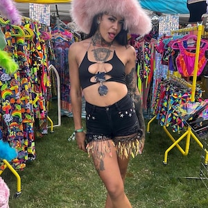 May include: A person wearing a black halter top and black denim shorts with star and fringe details. The person is wearing a large pink feathered hat and black boots. The background includes racks of colorful clothing and accessories.