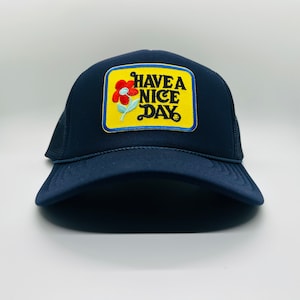 May include: A navy blue trucker hat with a yellow patch that says "HAVE A NICE DAY" with a red and white flower graphic.