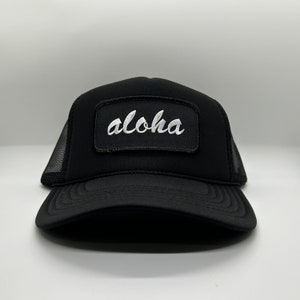 May include: A black mesh trucker hat with a black patch that says "Aloha" in white cursive lettering.