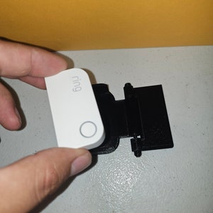 Op de afbeelding: Een wit Ring slimme thuistoestel met het woord "Ring" erop gedrukt. Het apparaat wordt in de hand gehouden en bevindt zich naast een zwarte kunststof montagebeugel.