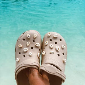 Puede incluir: Un par de zuecos beige decorados con pequeñas conchas blancas. Los zuecos se sostienen en una mano contra un fondo de piscina azul.