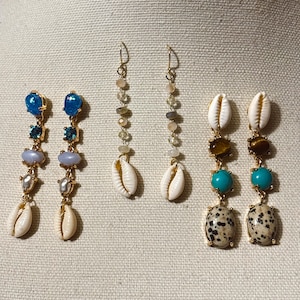 May include: Three pairs of drop earrings featuring gold-tone metal chains. The first pair has blue stones, light purple beads, and white cowrie shells. The second pair features small neutral beads and white cowrie shells. The third pair has brown, turquoise, and spotted stones with white cowrie shells.