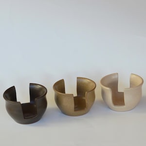 May include: Three ceramic cup holders in different shades of brown. The cup holders are designed to hold a cup or mug in the centre.