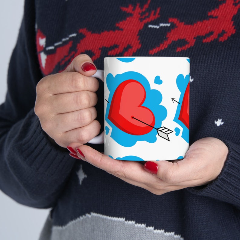 May include: White ceramic mug featuring a red heart design with an arrow through it. The heart is surrounded by blue clouds and smaller hearts.