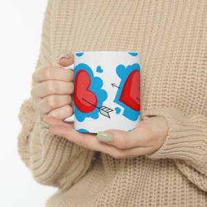 May include: White ceramic mug featuring a red heart design. The hearts are outlined in blue and have a red arrow through them. The mug is being held by a person's hands.