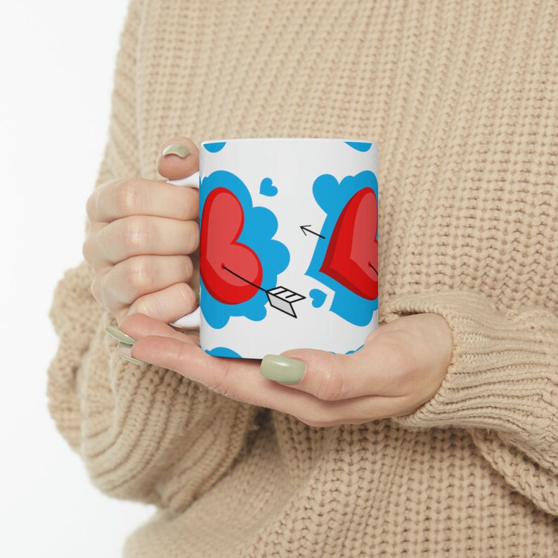 May include: White ceramic mug featuring a red heart design. The hearts are outlined in blue and have a red arrow through them. The mug is being held by a person's hands.