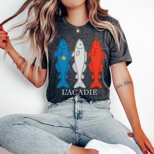 Peut inclure: T-shirt gris foncé avec trois poissons stylisés en bleu, blanc et rouge, avec le texte "L'ACADIE" en dessous. Le poisson bleu a une étoile jaune. C'est un vêtement décontracté.