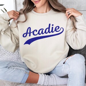 May include: A cream-coloured sweatshirt featuring the word "Acadie" in blue script. The word has a blue shadow, and the sweatshirt is a crew neck style. The person is wearing light blue jeans.