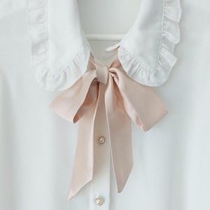 May include: A white blouse with a ruffled collar and a pink satin bow tie. The blouse has pearl buttons.