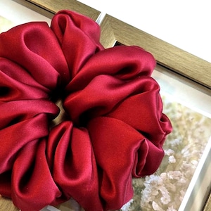 May include: A deep red, satin-finish hair scrunchie with a gathered, voluminous design. The fabric has a smooth, reflective texture with soft folds throughout the circular shape, creating a dense and pleated appearance for this hair accessory.