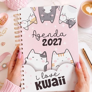 Könnte beinhalten: Ein spiralgebundener 2027-Terminplaner mit hellrosa Einband, der mit Cartoon-Katzen sowie dem Text Agenda 2027 und i love KWAII bedruckt ist. Der Einband hat einen weißen, wolkenförmigen unteren Bereich mit rosa Sternakzenten und eine goldene Spirale.