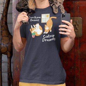 May include: A dark grey t-shirt with a graphic of two orange cats. The text on the shirt reads "Sunshine Beams" and "Catnip Dreams".