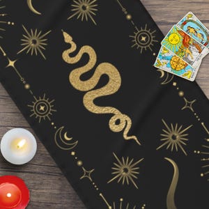 May include: Black fabric with a gold snake design and a repeating pattern of stars, moons, and suns. The fabric is on a wooden surface with three tarot cards.