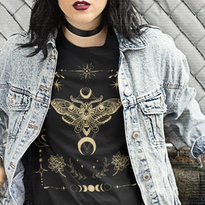 May include: Black t-shirt with a gold moth and celestial design. The graphic includes a moth with a skull, crescent moons, stars, and phases of the moon. The shirt is worn with a denim jacket.