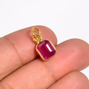 May include: A gold-colored pendant with a rectangular, deep red gemstone. The gemstone is set within a gold bezel and has a polished surface. The pendant has a small loop for attaching to a chain or necklace.