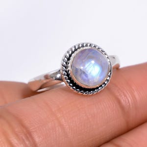 May include: A silver ring with a round, iridescent moonstone center. The moonstone is surrounded by a twisted rope-like border. The ring is set on a simple band.