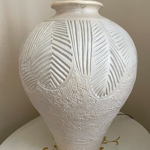 May include: White ceramic table lamp with a textured, carved design of leaves. The lamp has a white shade and a wooden base.