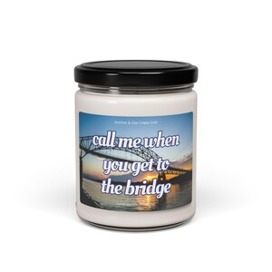 Op de afbeelding: Een witte kaars in een glazen pot met een zwart deksel. Het etiket toont een brug bij zonsondergang en de tekst "call me when you get to the bridge". De tekst "Anchor & Oar Cape Cod" is ook zichtbaar.