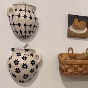 May include: A kitchen scene featuring decorative wall-mounted ceramic vases with black and white patterns. A small painting of pears hangs above a woven basket filled with potatoes. A basket of bananas and an air fryer are on the countertop.