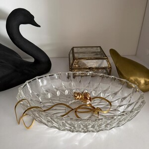 May include: A clear, circular glass bowl with a faceted, diamond-cut pattern on the exterior. Inside the bowl rests a thin, gold-toned metal chain necklace. The bowl is positioned on a flat surface near a black swan figurine and a small glass box.